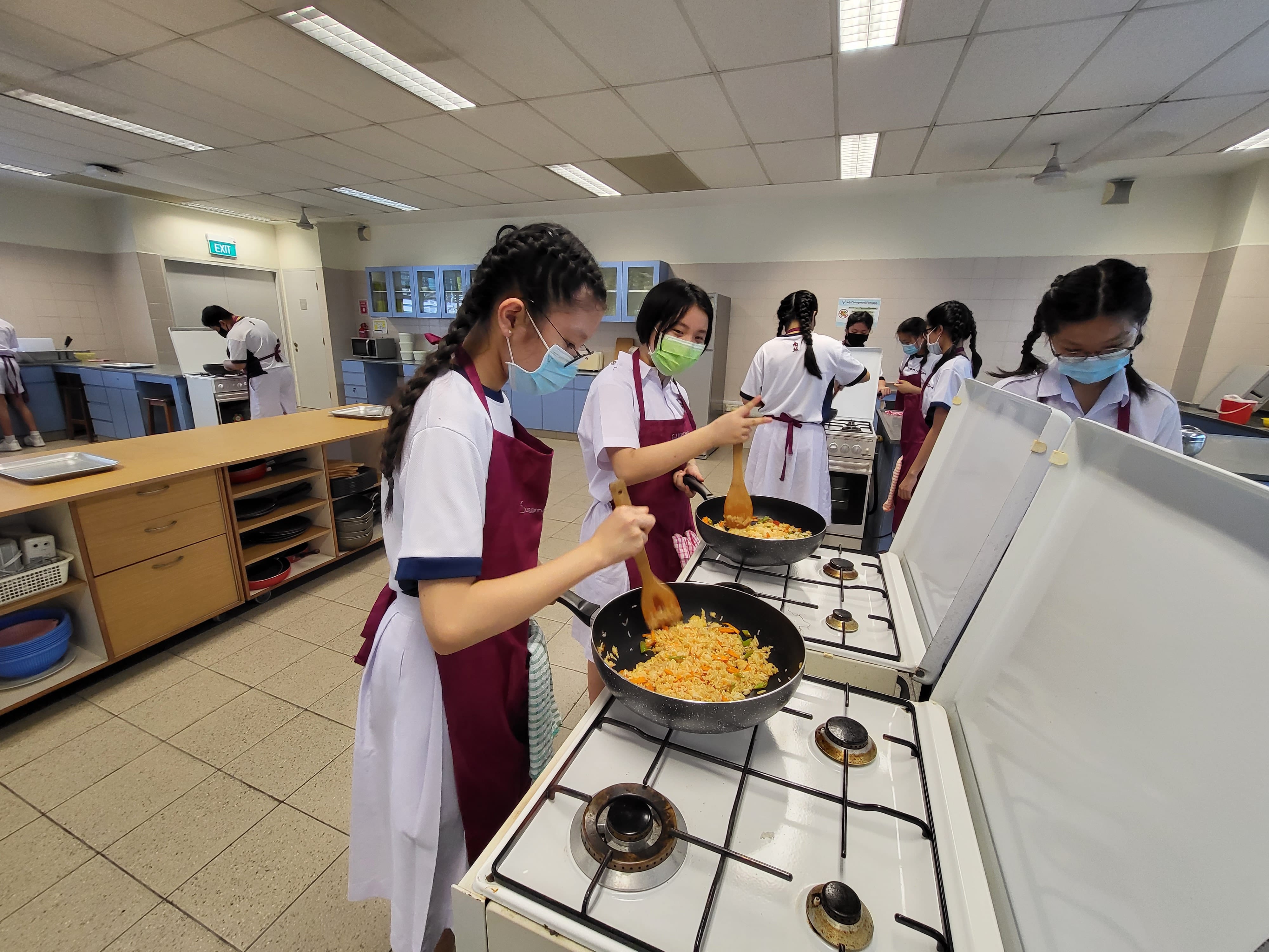 Students in aprons and face masks stir-fry rice in pans on a gas stove in a school cooking classroom.